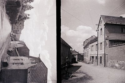 This evocative black-and-white image captures a narrow street scene, possibly in a European alpine village. Stone buildings w...