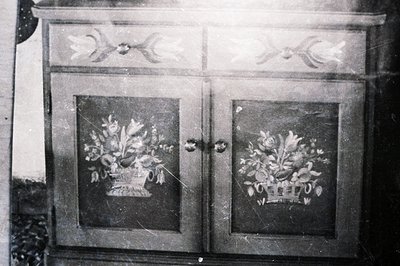 Detailed, grayscale image of antique furniture with decorative floral motifs painted on two cabinet doors. The cabinet featur...