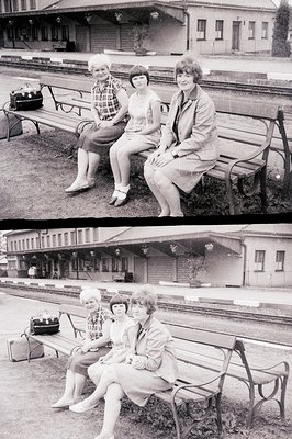 Three women sit on a bench outside a brick station building, likely awaiting or departing a train. Fashion suggests a 1960s o...