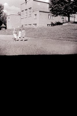 Two young girls in dresses stand before a modernist, two-story building with large windows. A grassy slope rises to the struc...