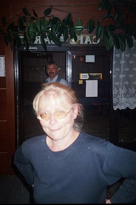 Close-up portrait of a woman with short, reddish-blonde hair and glasses, positioned outside a building with decorative folia...