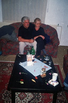 Two men relax on a plush, floral-patterned sofa in a casual domestic setting. A low, dark coffee table displays remnants of a...