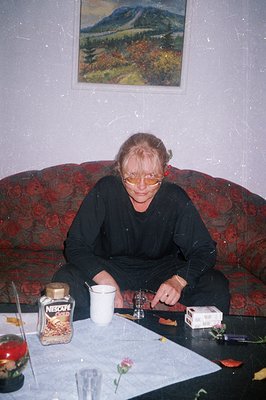 A seated woman with fair hair rests on a floral-patterned sofa. A table with coffee, a cup, and smoking paraphernalia is clos...