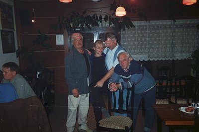 Four men appear in a dimly lit indoor scene, possibly a restaurant or banquet hall. Three of the men are in the foreground; o...