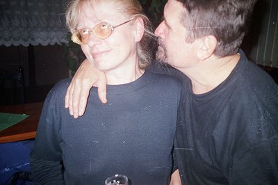 A man and woman sit close together, the man’s arm around the woman's shoulder. She wears glasses and a black turtleneck. The ...