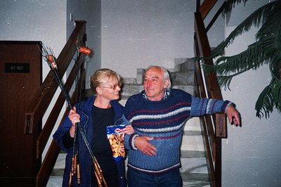 A smiling couple stands on a staircase, carrying bamboo skewers loaded with grilled sausage. Man in a striped sweater and wom...