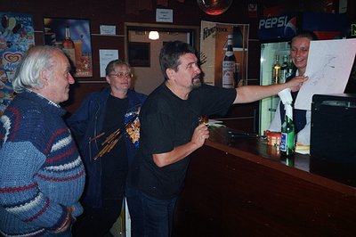 A group gathers at a bar, a man presenting a document or drawing. The decor suggests a casual, possibly rural, establishment....