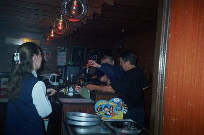 A photo shows a DJ at a mixing console with multiple turntables in a dimly-lit, wood-paneled club. A young woman watches from...