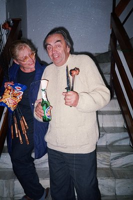 Two men stand on a staircase, one in a dark jacket, the other in a cream-colored sweater. The first carries a bag of chips an...