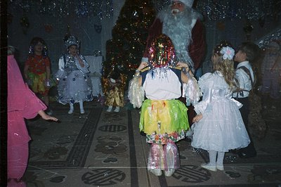 Children in costumes, including a young girl adorned with long, colorful streamers, gather around a figure presumed to be San...