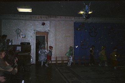 Children in costumes perform on a stage adorned with a blue backdrop and festive decorations. A piano sits to the left, and a...