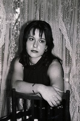 A young woman sits on a dark wooden chair, her arms crossed. She wears a sleeveless top and has short, dark hair with bangs. ...