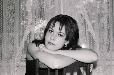 Striking portrait of a young woman with dark hair and bangs, arms crossed, leaning against a chair. Delicate lace curtains pr...