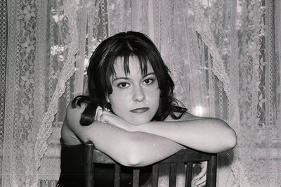A young woman with dark, shoulder-length hair and bangs sits before a lace-curtained window, arms crossed over a wooden chair...