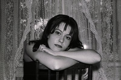 A young woman with dark hair and bangs leans forward, arms crossed on a dark wooden frame, likely a window or doorway. A lace...