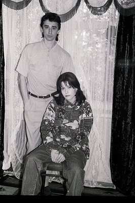 A young couple poses for a formal portrait. The woman sits on a chair, wearing a patterned sweater and trousers, while the ma...