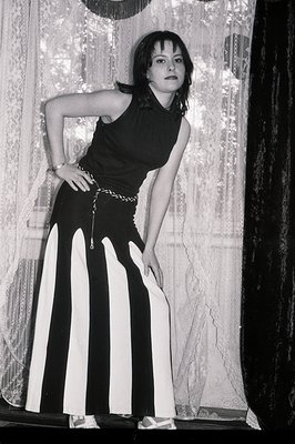 Young woman posed indoors, wearing a black halter top, wide-striped, floor-length trousers, and sandals. She leans against dr...