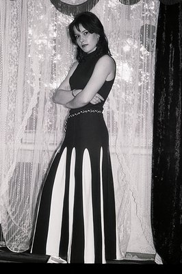 Striking portrait of a woman in a black and white, full-length, pleated dress. She stands with arms crossed, looking directly...