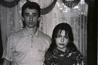 A monochrome portrait depicts a couple, likely in the 1980s. The man wears a polo shirt, arm around the woman, who is adorned...