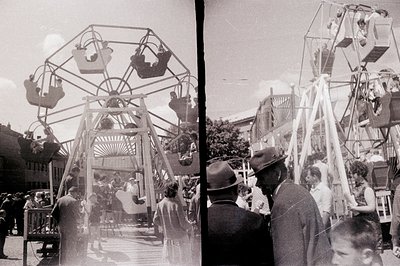 A vintage Ferris wheel dominates this view, transporting passengers above a crowded scene. People in 1960s attire—suits, dres...