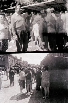 A black and white street scene shows a crowd of people clustered under a roofed area and along a paved road, likely in Easter...