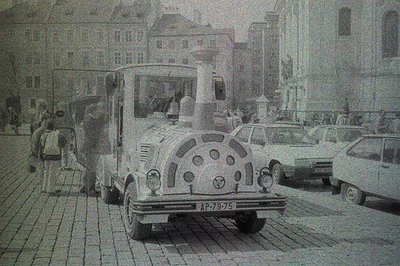 Black & white photograph of a vintage bus in a European city square. Architectural details suggest a historic urban setting, ...