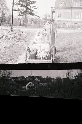 A woman in a long coat pushes a wicker baby carriage along a tree-lined road. A young child is visible within the carriage. A...