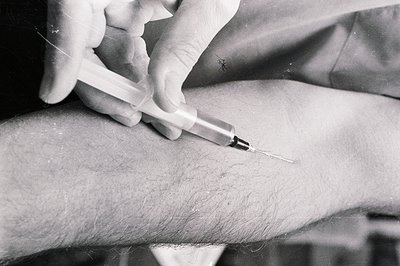 Close-up, black and white image depicts a gloved hand administering an injection into a hairy arm. The syringe needle is visi...