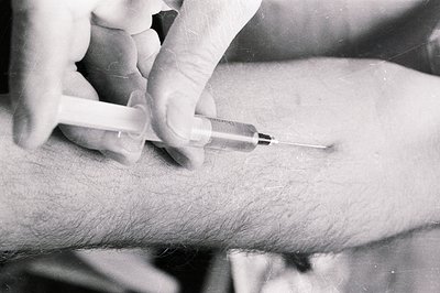 Close-up of a hand administering an injection into the arm. A syringe, labeled "Hikari," is visible. Grainy black and white f...