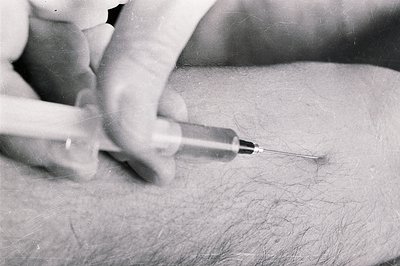 Close-up, black and white image shows a syringe poised to inject into a hairy arm. Appears to be a medical or scientific cont...