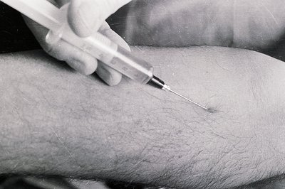 Close-up, black and white photo shows a hand administering an injection into a visibly hairy arm. The syringe needle is inser...