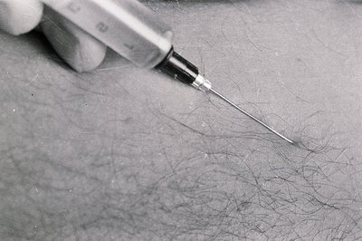 Close-up, monochrome image of a hypodermic needle poised above a textured surface covered in dark hair. The syringe’s marking...