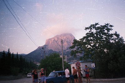 A group gathers around a vintage blue taxi, facing a dramatic, steep mountain under a dusky sky. Buildings and lush greenery ...