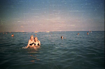 Two young women in swimwear smile while wading in a turquoise sea. Background shows numerous bathers and distant boats under ...