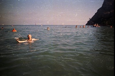 A wide, grainy photograph depicts multiple figures swimming in a calm, teal-colored sea. A single swimmer, centered and facin...