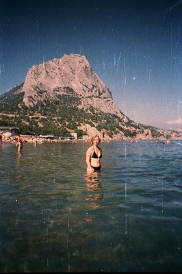 A young woman stands waist-deep in shallow, turquoise water, gazing towards a large, rocky mountain. A few other figures are ...