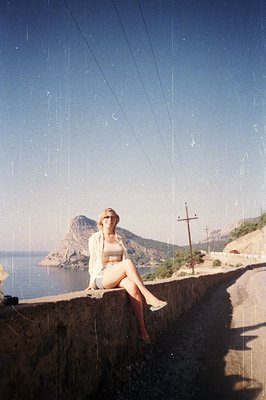 A young woman seated on a low stone retaining wall overlooks a coastal landscape. She wears a light-colored jacket, patterned...