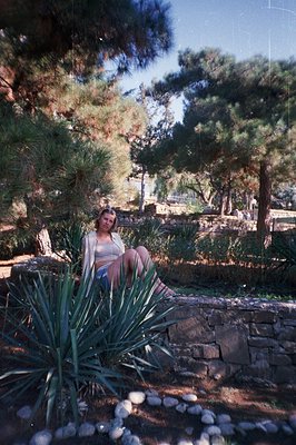 A young woman sits on a stone wall amidst lush greenery. The scene features a low stone wall covered in gravel and a large ag...