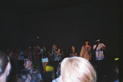 A concert scene with a band performing on stage. Visible are a vocalist in a patterned top and skirt, a guitarist, and a keyb...