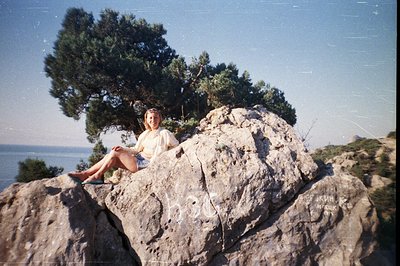 A woman sits atop a rocky outcrop overlooking the coastline. She wears a loose-fitting top and shorts, appearing relaxed. Den...