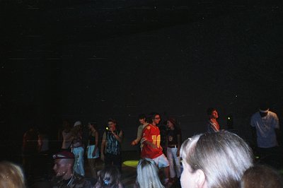 A group of young people gather outdoors, illuminated by stage lighting. Mixed clothing styles suggest a late 1990s/early 2000...