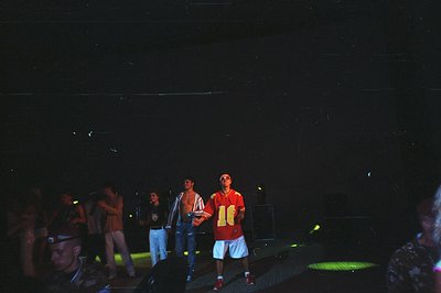 Group of men onstage, likely a performance or event. Stage lighting illuminates them; one wears a sports jersey and shorts. D...