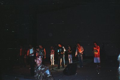 A group of young people gathered on a dimly lit stage, possibly a concert or performance. Notable for the individual in milit...