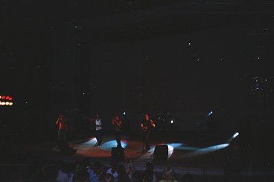 Concert stage captures four performers in the spotlight, silhouetted against a backdrop of stage lighting. A blurred crowd su...