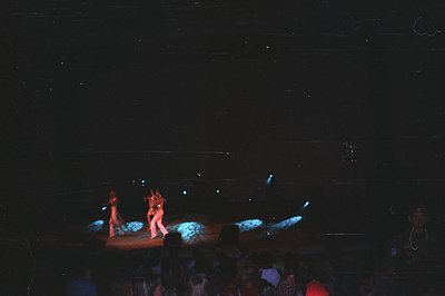 Three dancers perform on a stage illuminated with blue and red stage lights. A seated audience fills the foreground, blurred ...