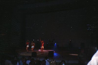 Three performers in matching swimwear and short, athletic shorts dance on a dimly lit stage. Professional lighting, including...