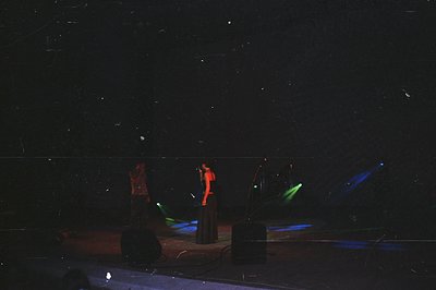 Stage performance featuring three figures in long, dark garments illuminated by red and green spotlights. Speakers and indist...