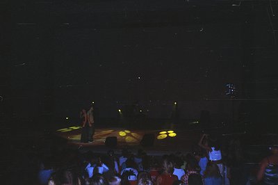 A concert scene shows two figures on stage illuminated by spotlights. A seated audience fills the foreground, creating a blur...