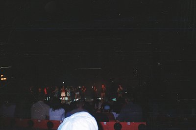 A crowd gathers before a brightly lit stage, potentially a concert or performance. Visible figures on stage suggest a musical...