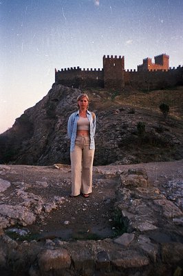 A woman stands before a fortified hilltop structure, likely a castle or fortress. She wears a cropped top & relaxed-fit trous...